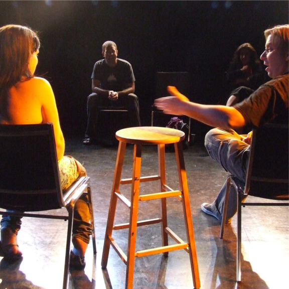 a group of people sitting around a wooden stool in drama studio