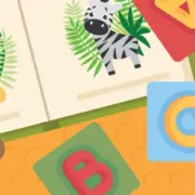 Illustration of a children's book open to pages showing a lion and a zebra, with a teddy bear and alphabet blocks nearby on a patterned background.