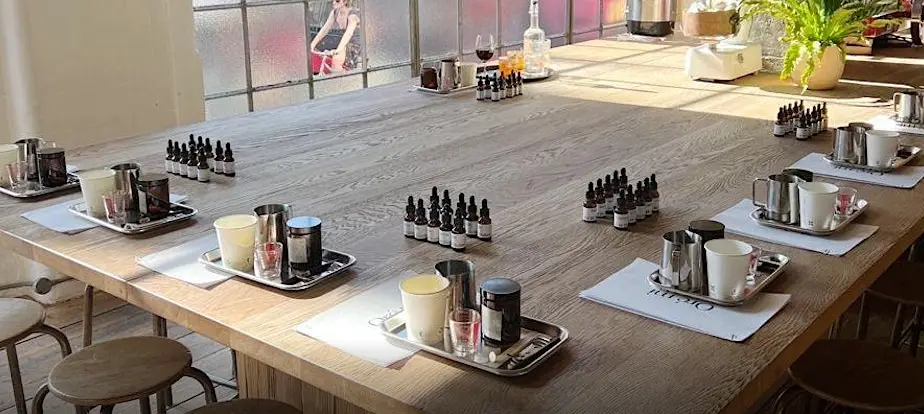 A large wooden table set up with trays containing candles, essential oil bottles, and other crafting supplies in a well-lit room.