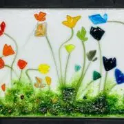 A rectangular glass artwork features colorful abstract flowers of various shapes and sizes against a white background, with green textured grass along the bottom.