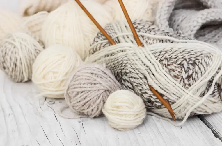 Several balls of yarn in various shades of white, cream, and gray are piled together on a wooden surface, with two wooden knitting needles resting on top.