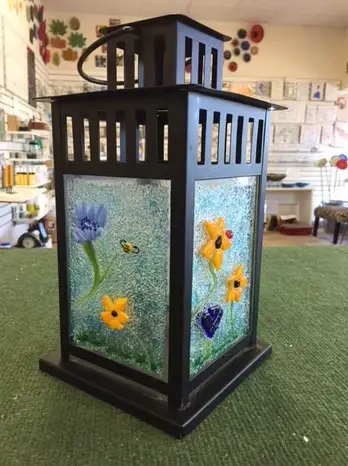 A black metal lantern with a glass panel featuring a painted design of flowers and a butterfly, placed on a green surface inside a room with various artwork displayed on the walls.