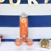 A table with a blue and white striped backdrop displays a large glass dispenser filled with a pink drink, four glasses, and trays of snacks. Two vases with pink flowers flank the setup.