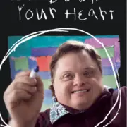 A person with a broad smile holds a marker, extending it towards the camera. Behind them is a colorful abstract painting. Text above reads "Lay Down Your Heart.