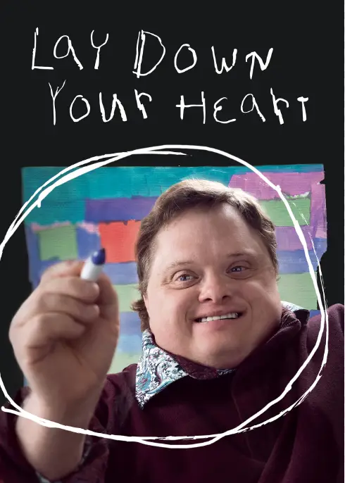 A person with a broad smile holds a marker, extending it towards the camera. Behind them is a colorful abstract painting. Text above reads "Lay Down Your Heart.